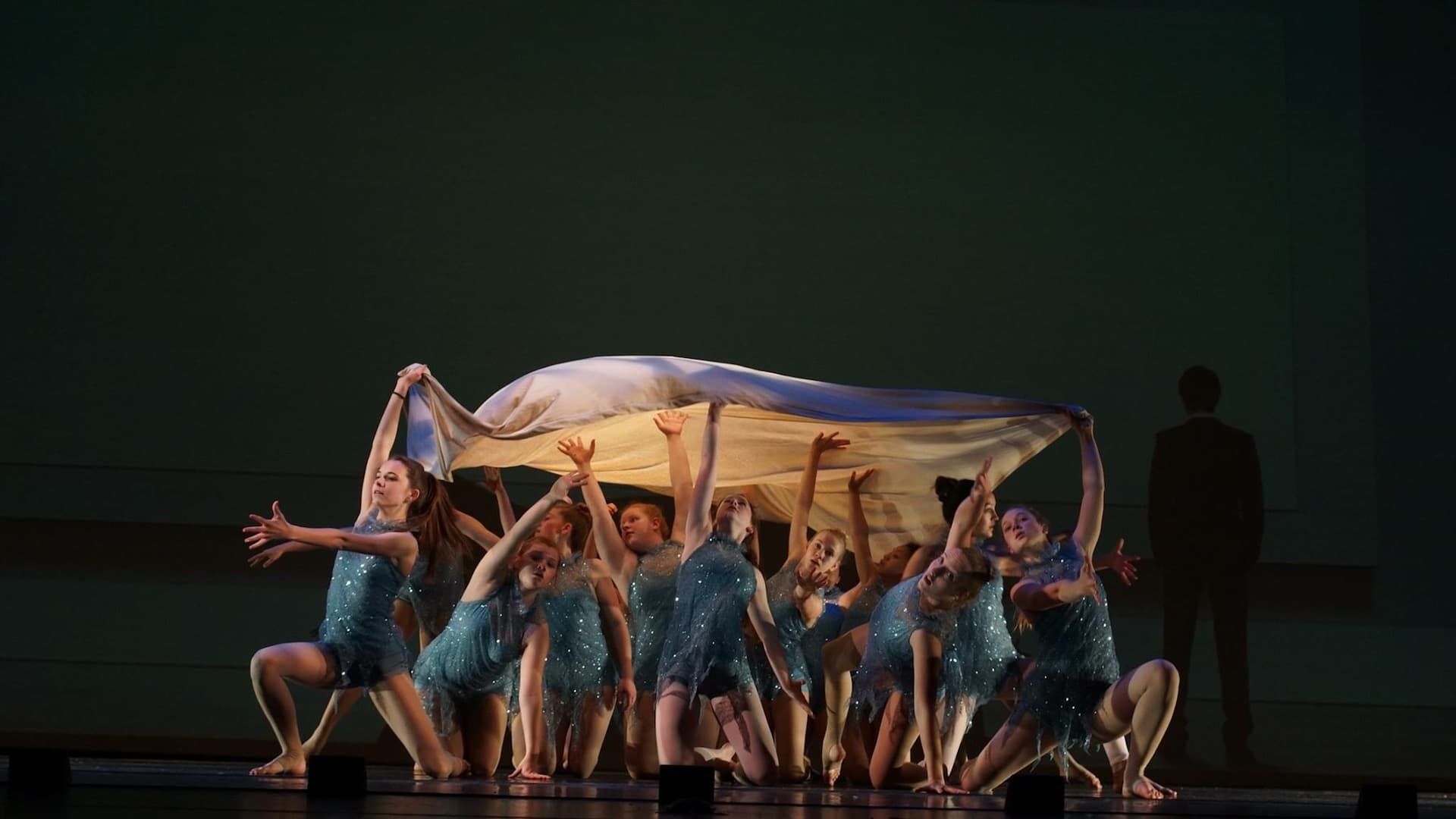 Dancers in sparkling teal costumes performing on stage under a flowing fabric prop