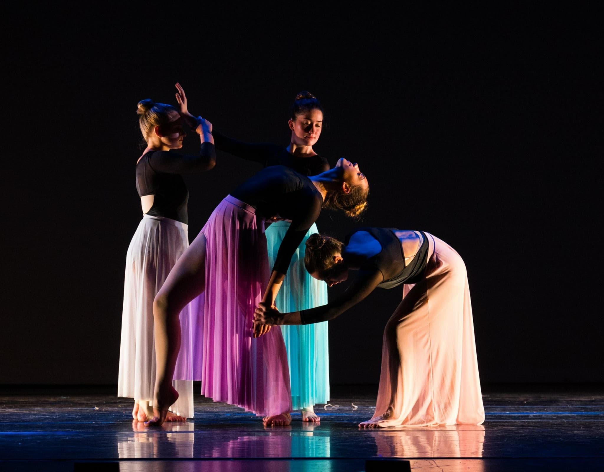 Four contemporary dancers in flowing skirts lifting and supporting each other on a dark stage